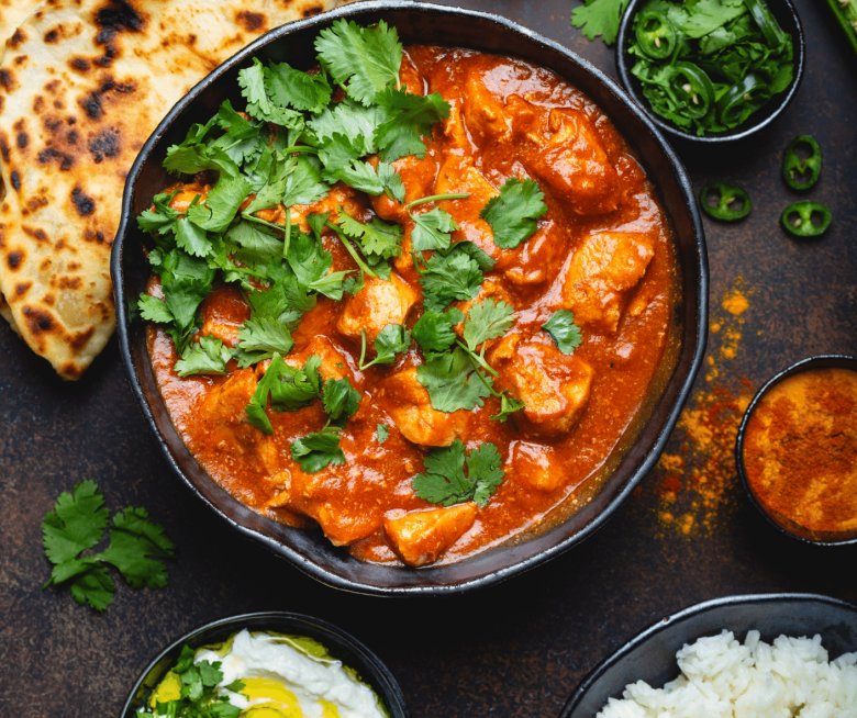Chicken Tikka Masala in einem Tontopf mit Koriander garniert, daneben Schälchen mit grünen Chilis und Joghurt