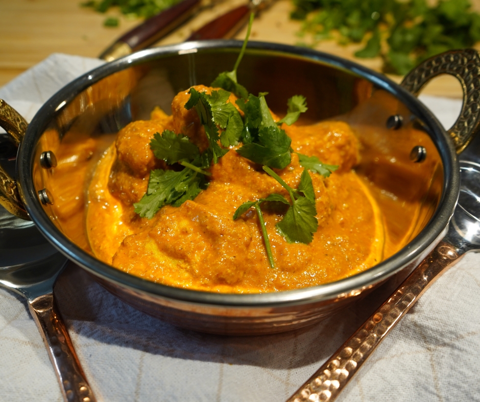 Leuchtend oranges, cremiges Butter Chicken in einer Kupferschale serviert und mit Korianderblättern garniert.