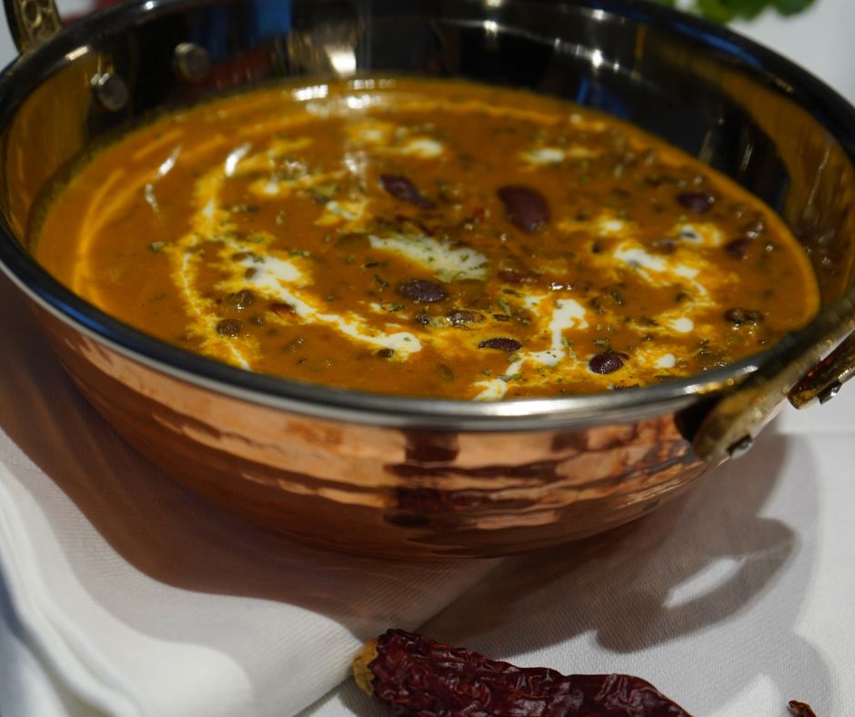 Dal Makhani in einer Kupferschale angerichtet und mit frischer Sahne und Methi-Blättern garniert. Vorne im Bild liegt eine getrocknete Kashmiri Chilischote.