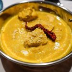Cremiges Cashew-Hähnchen-Curry angerichtet in einer Kupferschale auf einem weißen Tischtuch, garniert mit frischen Kokosflocken und einer roten Kashmiri-Chili. Im Hingergrund sieht man eine blaue Schale mit Koosflocken und einzelne Cashew-Nüsse.