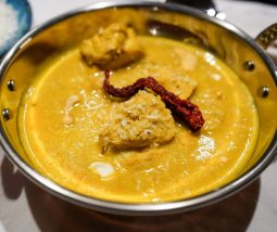 Cremiges Cashew-Hähnchen-Curry angerichtet in einer Kupferschale auf einem weißen Tischtuch, garniert mit frischen Kokosflocken und einer roten Kashmiri-Chili. Im Hingergrund sieht man eine blaue Schale mit Koosflocken und einzelne Cashew-Nüsse.