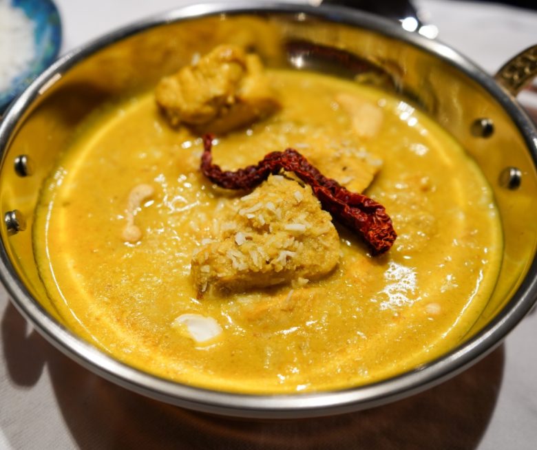 Cremiges Cashew-Hähnchen-Curry angerichtet in einer Kupferschale auf einem weißen Tischtuch, garniert mit frischen Kokosflocken und einer roten Kashmiri-Chili. Im Hingergrund sieht man eine blaue Schale mit Koosflocken und einzelne Cashew-Nüsse.