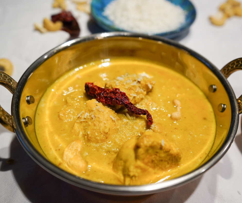Cremiges Cashew-Hähnchen-Curry angerichtet in einer Kupferschale auf einem weißen Tischtuch, garniert mit frischen Kokosflocken und einer roten Kashmiri-Chili. Im Hingergrund sieht man eine blaue Schale mit Koosflocken und einzelne Cashew-Nüsse.