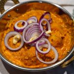 Leuchtend oranges, cremiges Dum Aloo - Indisches Kartoffelcurry, angerichtet in einer Kupferschale mit Cashew-Nüssen und frischen roten Zwiebelringen