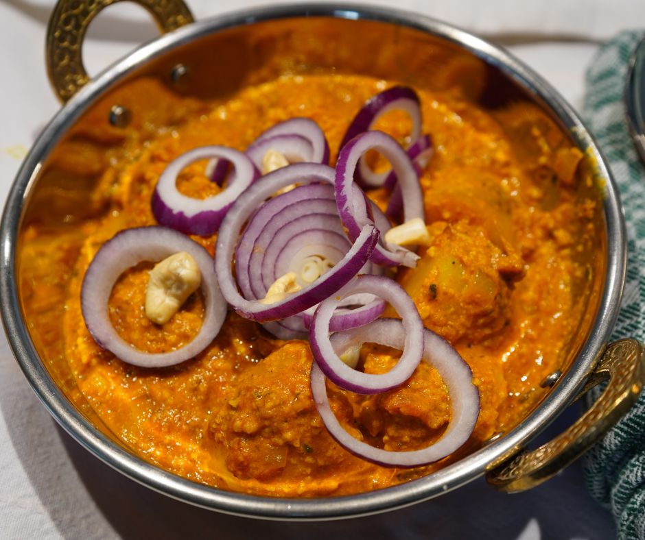 Leuchtend oranges, cremiges Dum Aloo - Indisches Kartoffelcurry, angerichtet in einer Kupferschale mit Cashew-Nüssen und frischen roten Zwiebelringen