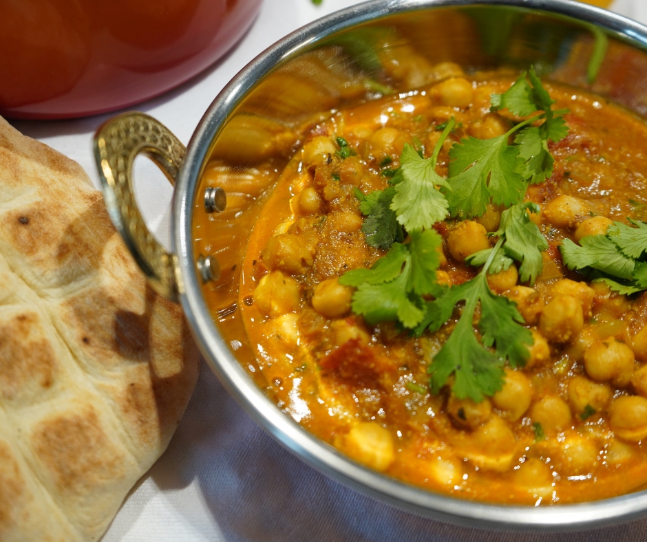 Du siehst eine Kupferschale mit leuchtend orangem Kichererbsen-Curry und Koriander. In einer roten Porzellan-Schale steht Basmati-Reis und links daneben ist ein Stück Fladenbrot zu sehen.