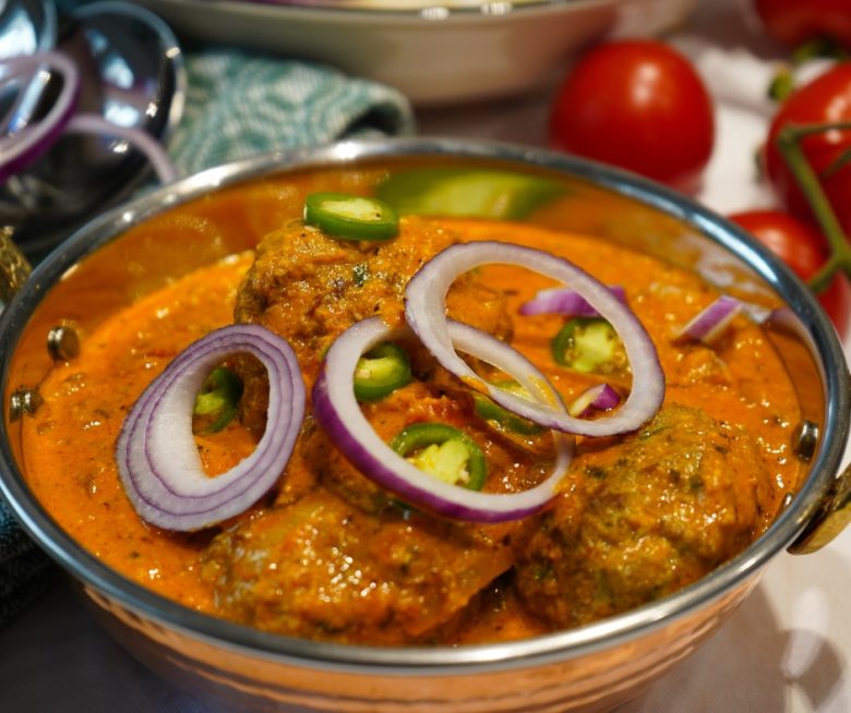 Indisches Kofta Curry mit Lamm in einer Kupferschale angerichtet und mit frischen, grünen Chilis und Scheiben roter Zwiebel garniert