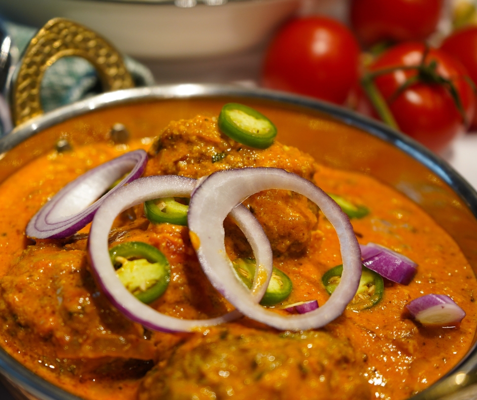 Indisches Kofta Curry mit Lamm in einer Kupferschale angerichtet und mit frischen, grünen Chilis und Scheiben roter Zwiebel garniert