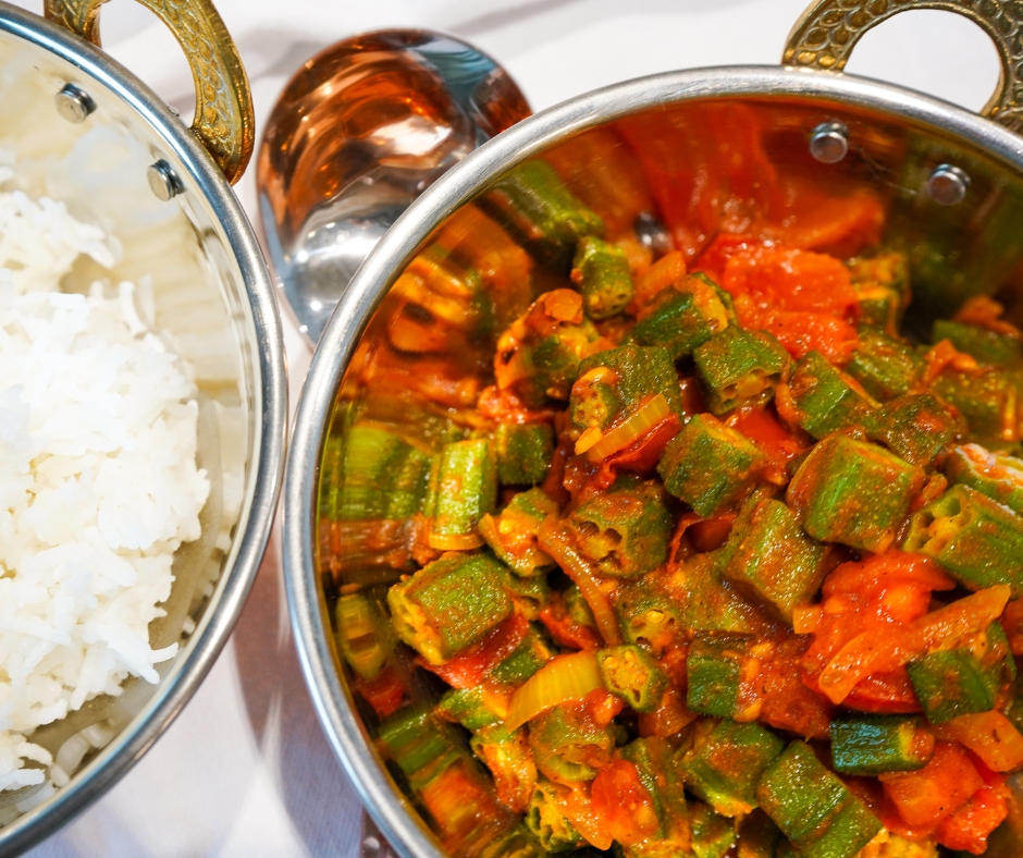 Leuchtend grün und rote Bhindi Masala mit Tomaten und Zwiebel angerichtet in einer Kupferschale, die neben einer zweiten Kupferschale voller Basmati-Reis steht.